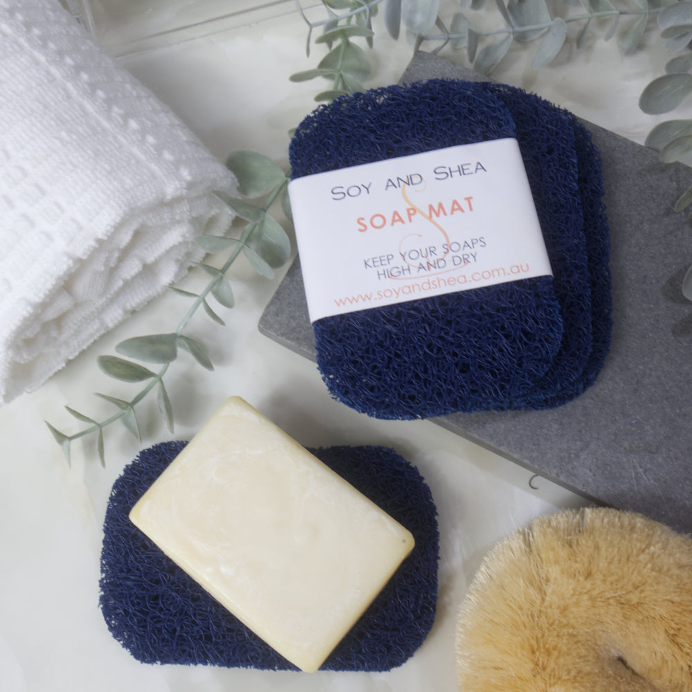 A flatlay image showing a collection of Dark Blue Soap mats, displayed on a bathroom countertop along with a white towel, body brush and greenery.  Three soap mats are stacked upon each other in a fan shape on a grey stone tray with the top one featuring a product label.  A fourth mat lies to the bottom of the image with a cream soap lying on it.