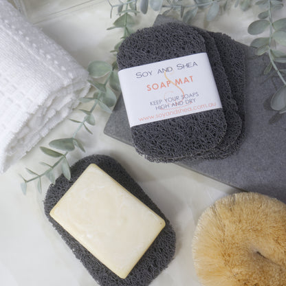 A flatlay image showing a collection of Dark Grey Soap mats, displayed on a bathroom countertop along with a white towel, body brush and greenery.  Three soap mats are stacked upon each other in a fan shape on a grey stone tray with the top one featuring a product label.  A fourth mat lies to the bottom of the image with a cream soap lying on it.