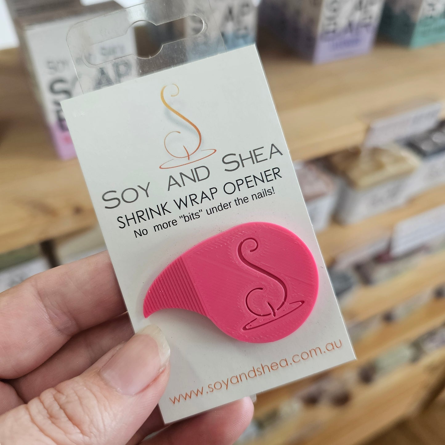 Pink Shrink wrap opener  with Soy and Shea logo on a white card held by a hand with a blurred background