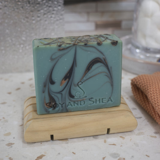 A green soap with black and gold swirls sits on a wooden dish with a blurred bathroom background