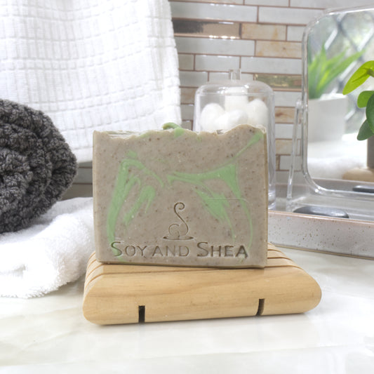 A bar of soap stands upright on a wooden soap dish with folded towels in the background. The soap has stone grey colouring with bright green swirls.  It has been stamped with the name Soy and Shea with a stylised S logo
