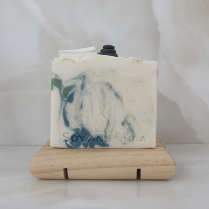 A bar of soap that is off white with dark green and gold swirls sits on a wooden soap dish surrounded by marble tiles. The top features embellishments nade of soap representing a stack of massage stones and a rolled up white towel.