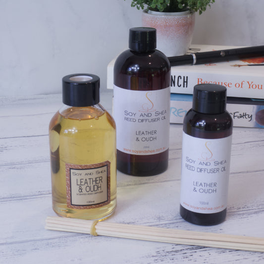 Three 'Leather and Oudh Reed Diffuser' bottles from Soy and Shea are elegantly placed on a white wooden table, filling the air with a rich, masculine scent. Two bottles are filled with reed diffuser oil, while one is topped with a smaller black cap. Reed sticks and stacked books subtly frame the scene, suggesting the allure of oudh wood.