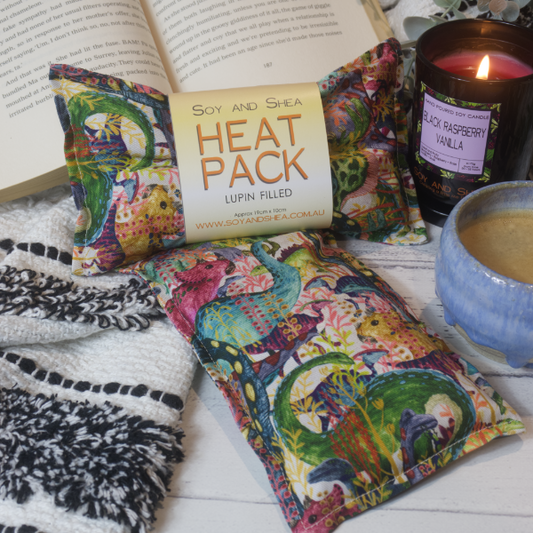 Two Mini Heat packs are stacked upon each other, one with a belly band label reading HEAT PACK LUPIN FIlled.   These are resting on a black and white blanket with open book, candle and coffee cup evoking a cosy feel.    The Heat packs show watercolour illustrations of a variety of dinosaur breeds surrounded by jungle vines.  