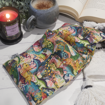 A small rectanglular Heat Pack rests on a wooden table top along with a black and white blanket, open book, burning candle and filled coffee cup evoking a cosy scene.  The Heat pack features many watercolour dinosaurs in earthy blue, yellow and green tones wondering through jungle vines.