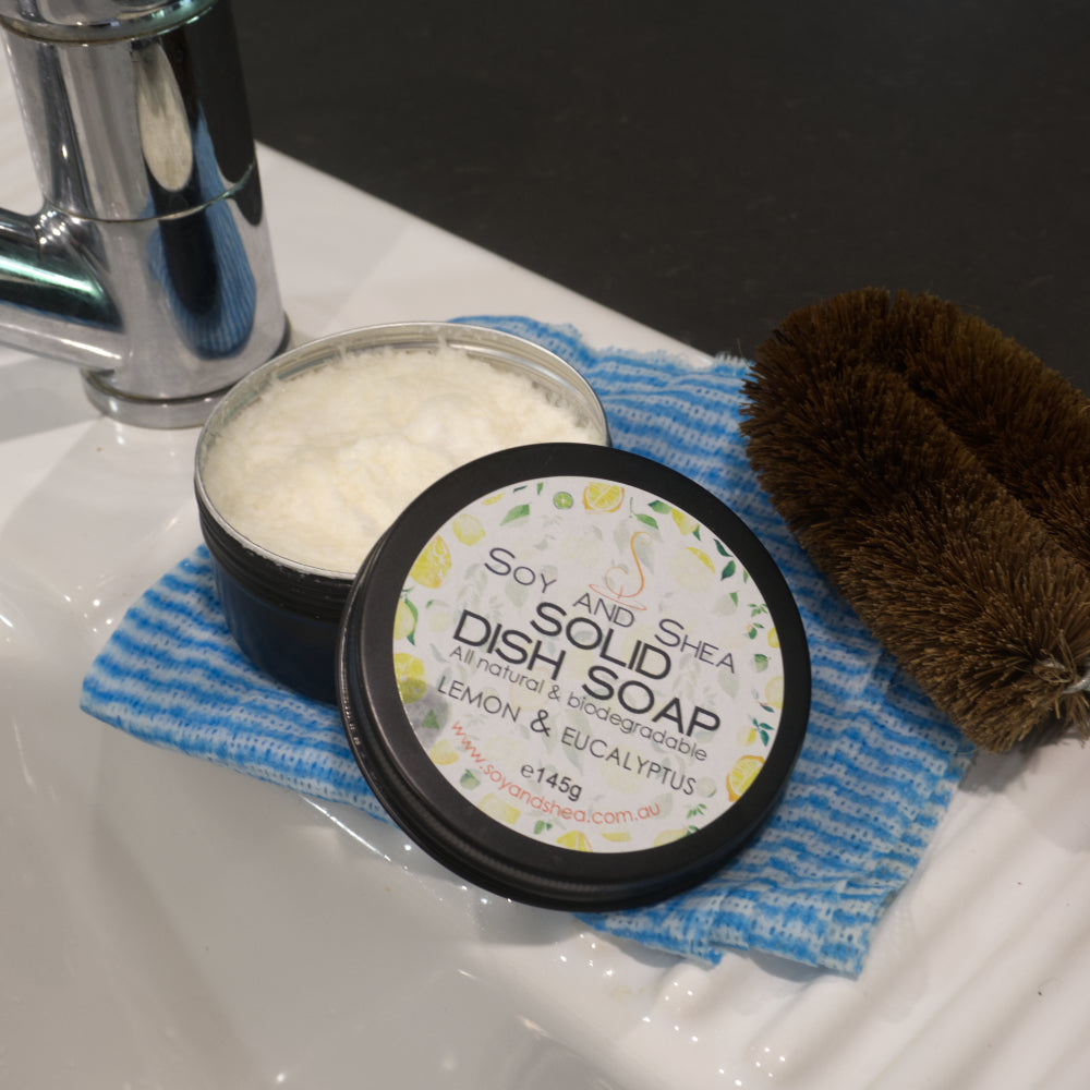 Solid dish soap in a container on a blue dish cloth with a scrub brush next to it on a sink.