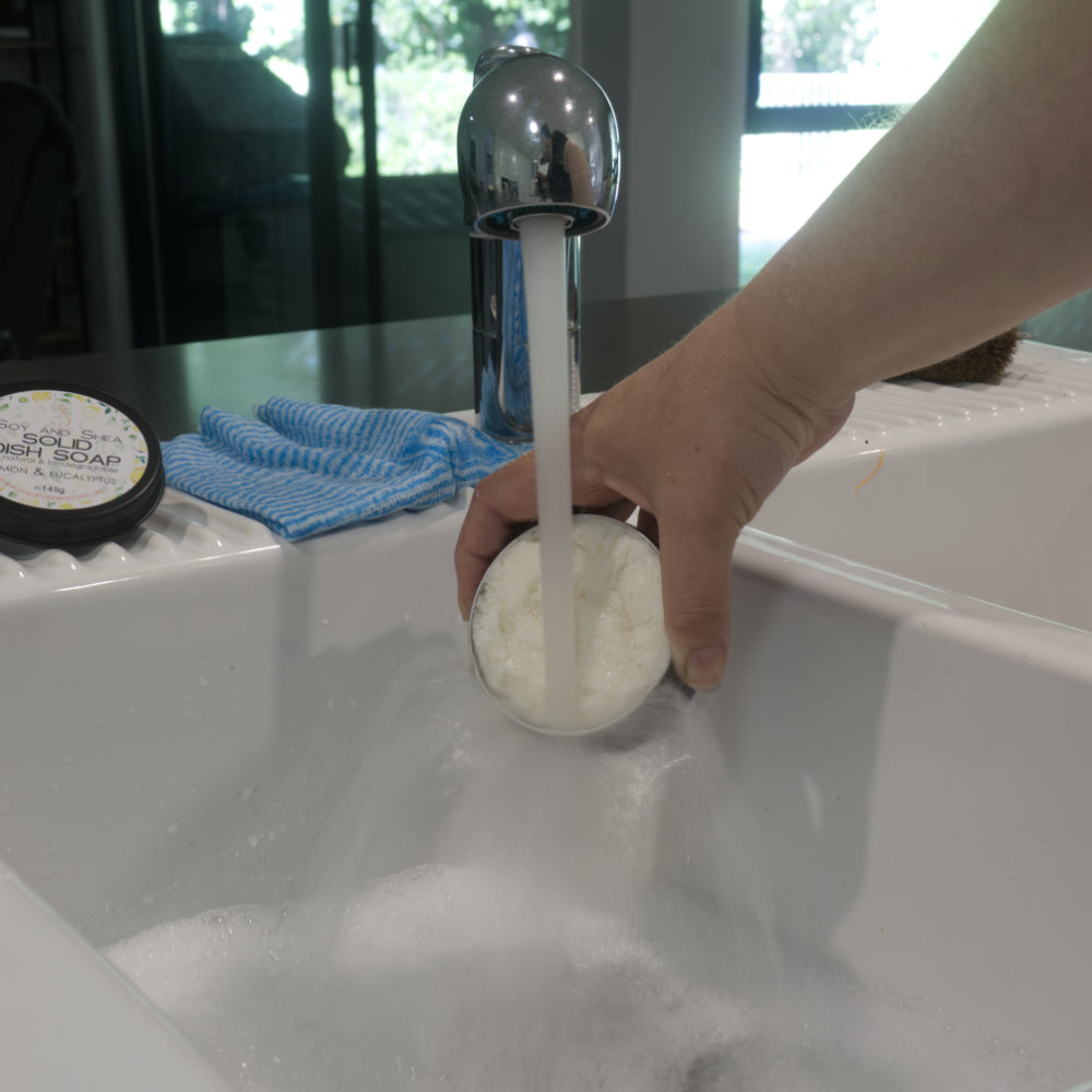 Hand holding a tin of Solid Dish Soap under water running into a sink with bubbles