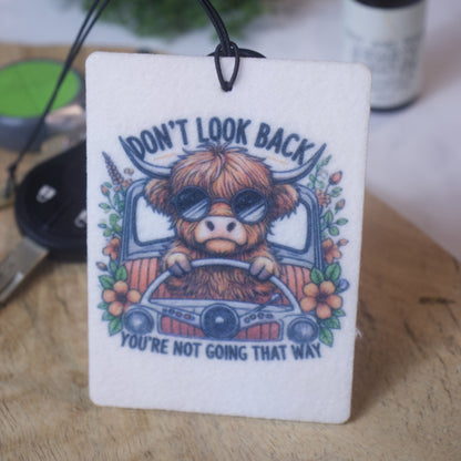 A car air freshener with an illustration of a highland cow wearing sunglasses behidn the wheel of a car, with text that reads 'DON'T LOOK BACK YOU'RE NOT GOING THAT WAY'.