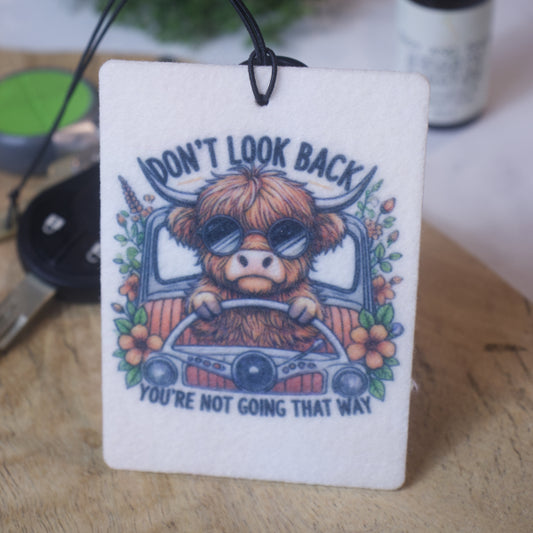 A car air freshener with an illustration of a highland cow wearing sunglasses behidn the wheel of a car, with text that reads 'DON'T LOOK BACK YOU'RE NOT GOING THAT WAY'.