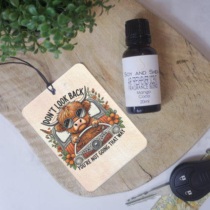 A rectangular car air freshener lies on a wooden surface next to a glass bottle of Air Freshener Scent and car key.  The Air Freshener has a cream background with the image of a highland cow behind the wheel of an antique car surrounded by orange flowers.  The text DON'T LOOK BACK YOU'RE NOT GOING THAT WAY  is written above and below the scene