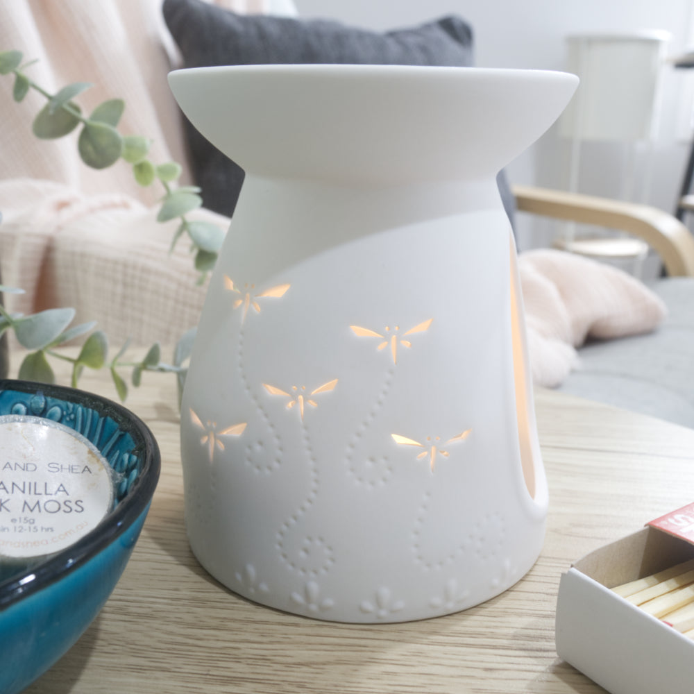 A matte white porcelain fragrance warmer sits on a light wooden table next to a blue bowl of wax melts and a box of matches. The warmer features embossed details at the base and lit dragonfly cutouts that glow warmly from an internal tealight candle. In the blurred background, a pink textured throw blanket and a dark grey cushion rest on a chair