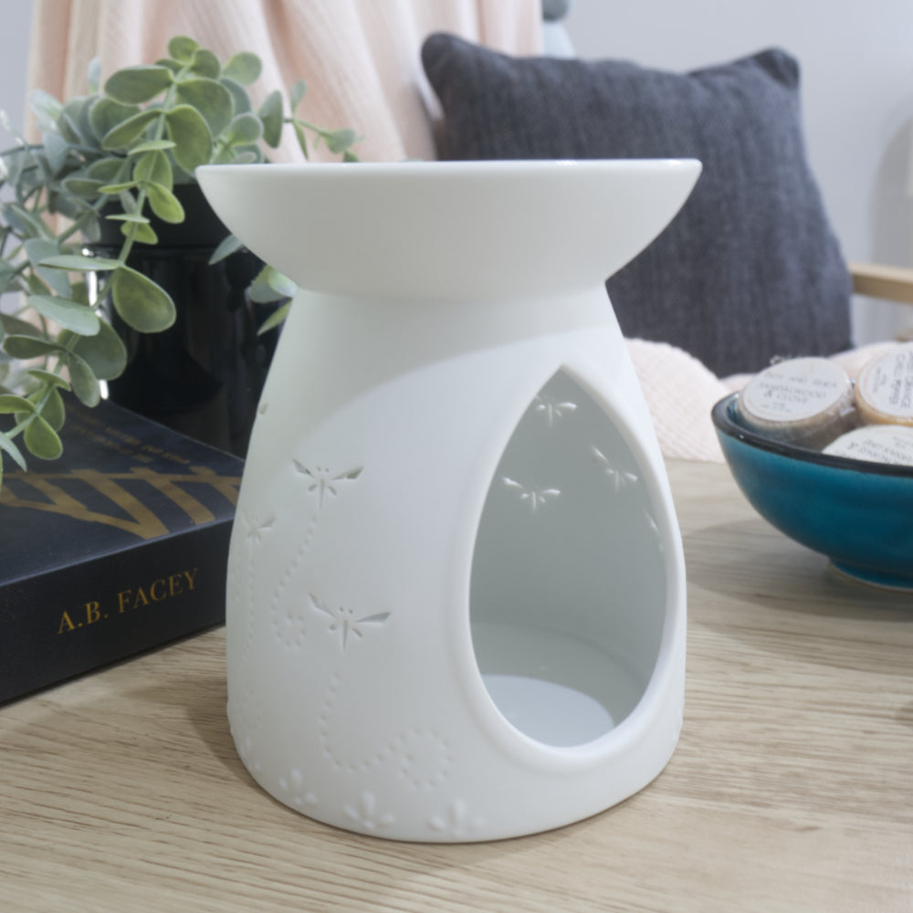 A matte white porcelain fragrance warmer sits on a light wooden table next to a blue bowl of wax melts and a box of matches. The image shows the warmer from the side showing large teardrop openings on the side of the warmer.