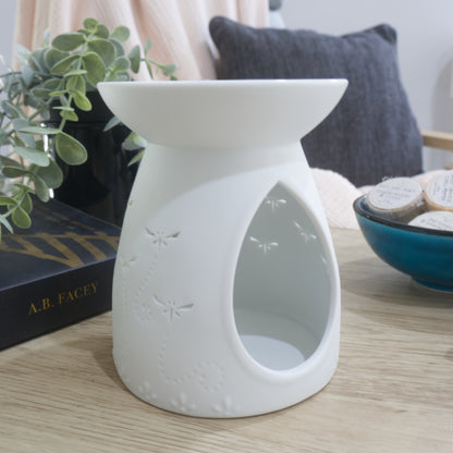 A matte white porcelain fragrance warmer sits on a light wooden table next to a blue bowl of wax melts and a box of matches. The image shows the warmer from the side showing large teardrop openings on the side of the warmer.
