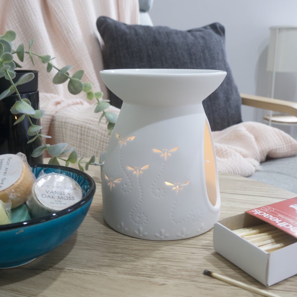 A matte white porcelain fragrance warmer sits on a light wooden table next to a blue bowl of wax melts and a box of matches. The warmer features embossed details at the base and lit dragonfly cutouts that glow warmly from an internal tealight candle. In the blurred background, a pink textured throw blanket and a dark grey cushion rest on a chair