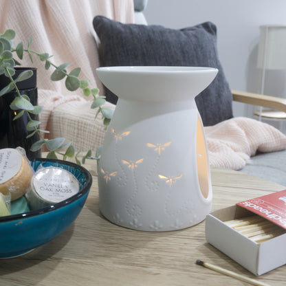 A matte white porcelain fragrance warmer sits on a light wooden table next to a blue bowl of wax melts and a box of matches. The warmer features embossed details at the base and lit dragonfly cutouts that glow warmly from an internal tealight candle. In the blurred background, a pink textured throw blanket and a dark grey cushion rest on a chair