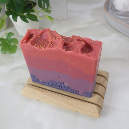 A bar of soaps stands on a wooden soap rack on a bathroom countertop along with a vine-like plant and stone tray.  The soap is pictured from above showing a red top with gold cellulous beads.   The soap has four jaggered layers of colour starting with dark then light purple.  Followed by light then bright red.  
