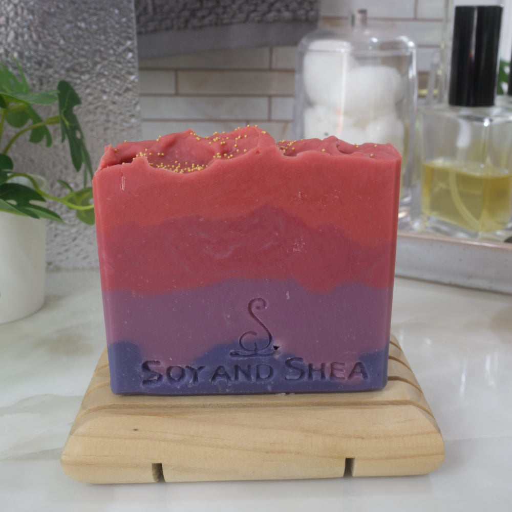 A bar of soaps stands on a wooden soap rack on a bathroom countertop.  Behind is various bathroom accessories including a towel, bottle of perfume and jar of cotton balls.  The soap has four jaggered layers of colour going from dark then light purple.  Followed by light then bright red.  The top of gold cellulous beads.