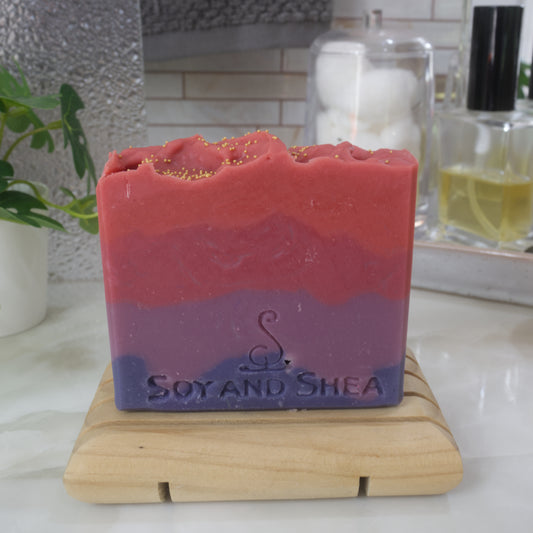A bar of soaps stands on a wooden soap rack on a bathroom countertop.  Behind is various bathroom accessories including a towel, bottle of perfume and jar of cotton balls.  The soap has four jaggered layers of colour going from dark then light purple.  Followed by light then bright red.  The top of gold cellulous beads.