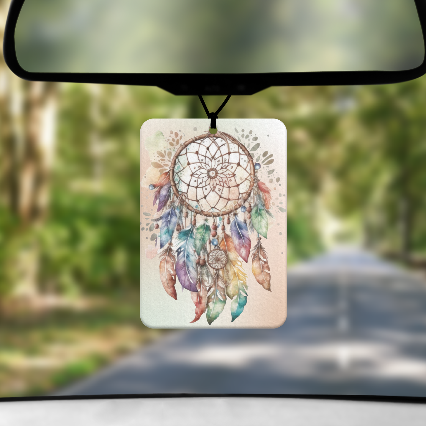 A mock up image showing a view through a car windscreen with a long tree lined road.  From the rearview mirror hands an air freshener with an image of dream catcher with feather tassels