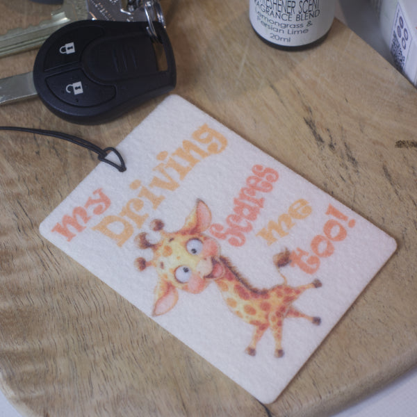  A rectangular car air freshener lies on a wooden surface beside a car key.   The air freshener features a cartoon style crazy giraffe surrounded by the words MY DRIVING SCARES ME TOO in shades of orange 
