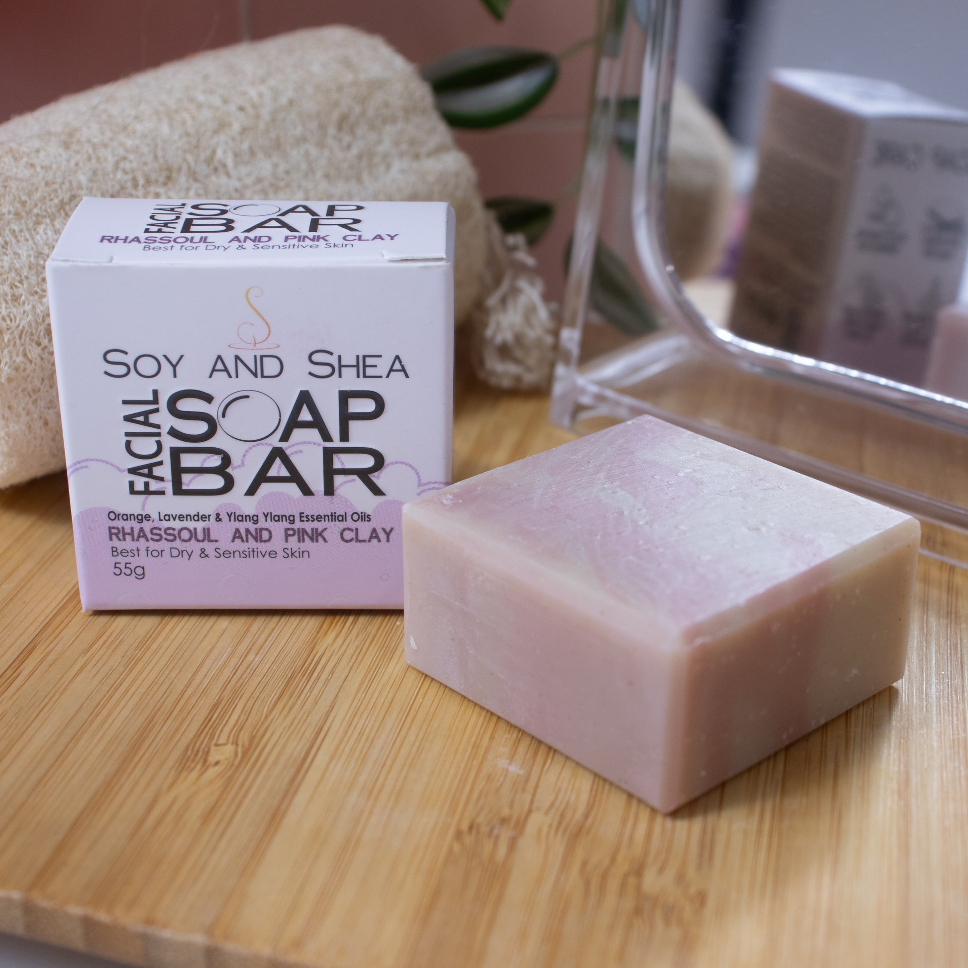 A small squarish soap bar with pale pink and white swirls lays flat on a wooden surface next to its packaging, a white box with a green border represent bubble foam.  In the background is a mirror, natural loofah and greenery.