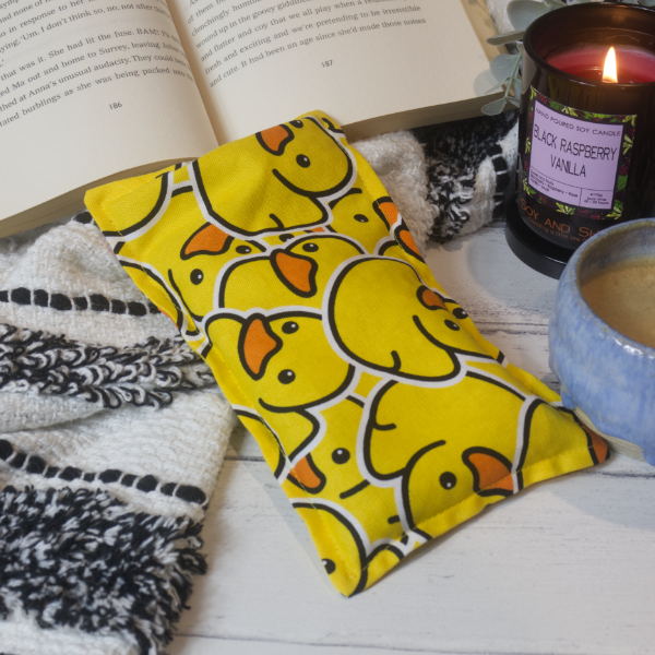 A mini rectangular heat pack rests against a black and white blanket and open book.  Next to it is a burning candle and filled coffee cup evoking a comfy scene.    The heat pack shows illustrations of lots of yellow rubber ducks stacked upon each other.