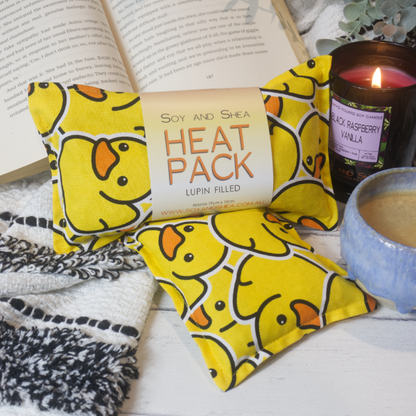 Two Mini Heat packs are stacked upon each other, one with a belly band label reading HEAT PACK LUPIN FIlled.   These are resting on a black and white blanket with open book, candle and coffee cup evoking a cosy feel.    The Heat packs feature bright yellow rubber ducks stacked upon each other.