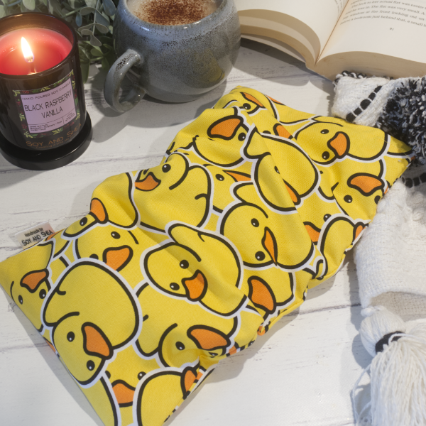 A small rectanglular Heat Pack rests on a wooden table top along with a black and white blanket, open book, burning candle and filled coffee cup evoking a cosy scene.  The Heat pack is a bright yellow featuring many rubber ducks stacked upon each other