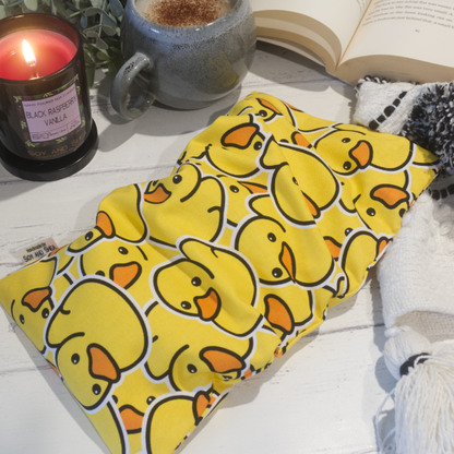 A small rectanglular Heat Pack rests on a wooden table top along with a black and white blanket, open book, burning candle and filled coffee cup evoking a cosy scene.  The Heat pack is a bright yellow featuring many rubber ducks stacked upon each other