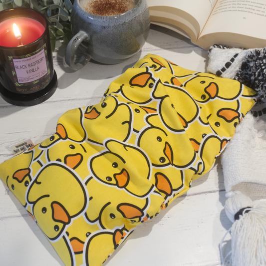 A small rectanglular Heat Pack rests on a wooden table top along with a black and white blanket, open book, burning candle and filled coffee cup evoking a cosy scene.  The Heat pack is a bright yellow featuring many rubber ducks stacked upon each other