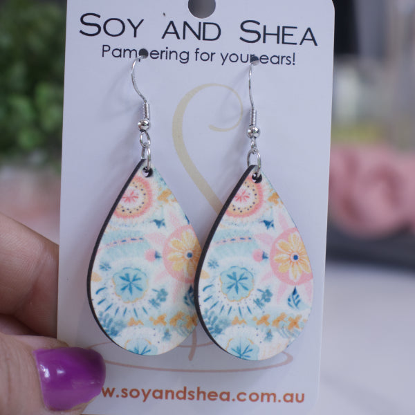 A hand holds a white card branded with SOY AND SHEA with a pair of earrings hanging on hooks.  The earrings are teardrop shaped featuring a pastel bohon floal design in shades of blue and pink.