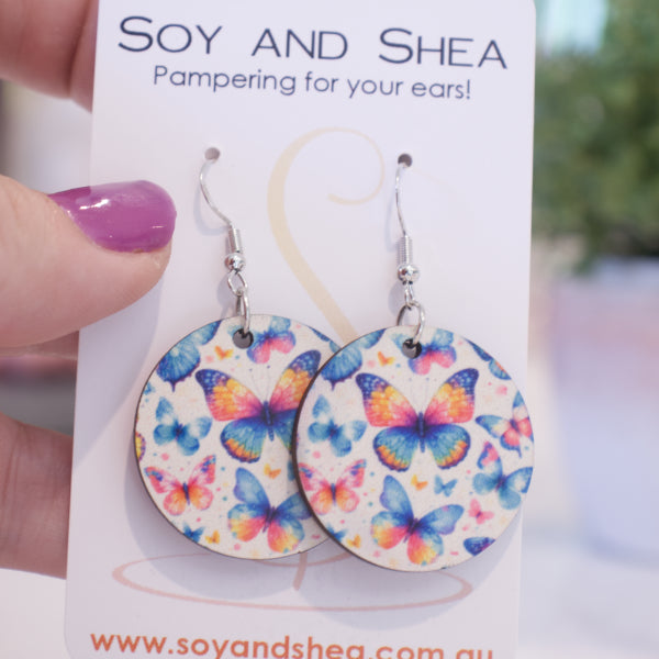 A hand holds a white card branded with SOY AND SHEA with a pair of earrings hanging on hooks.  The earrings are round with a white background covered in various sized butterflies in sahdes of blue, pink and yellow