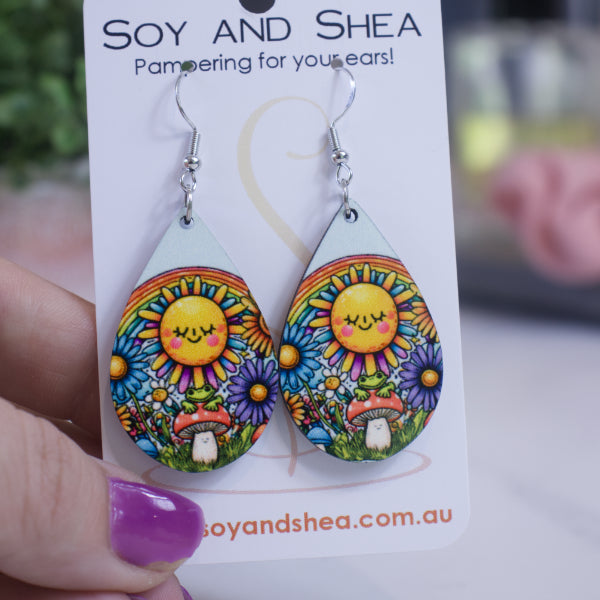 A hand holds a white card branded with SOY AND SHEA with a pair of earrings hanging on hooks.  The earrings are teardrop shaped featuring a large sunshine with kawai face in front of a rainbow.  Below is a toadstool with small green frog surrounded by wild flowers