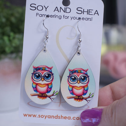 A hand holds a white card branded with SOY AND SHEA with a pair of earrings hanging on hooks.  The earrings are teardrop shaped with a light blue background.  The is a cartoon image of a pink owl with rainbow coloured wings and a head bandana sitting on a tree branch