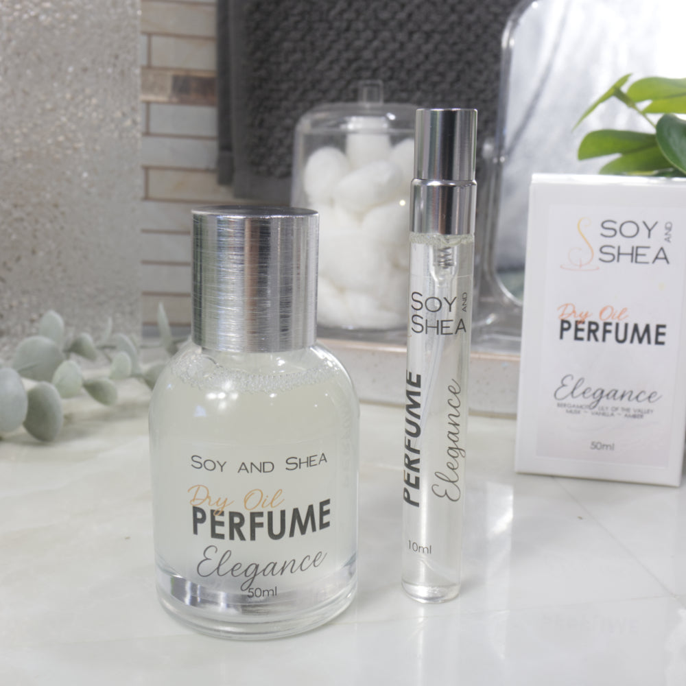 A selection of perfume bottles are presented on a bathroom countertop.  A clear, narrow, tall bottle labeled as 10ml with a silver cap stands beside a clear round, short bottle with silver lid labelled as 50ml.  Behind sits a white box with label identifying as Dry Oil Perfume Elegance.
