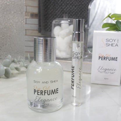 A selection of perfume bottles are presented on a bathroom countertop.  A clear, narrow, tall bottle labeled as 10ml with a silver cap stands beside a clear round, short bottle with silver lid labelled as 50ml.  Behind sits a white box with label identifying as Dry Oil Perfume Elegance.