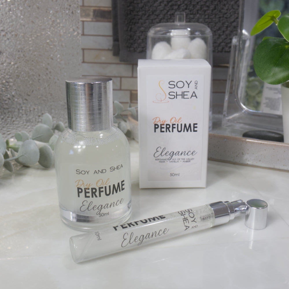 A selection of perfume bottles are presented on a bathroom countertop.  To the front is a clear, narrow, tall bottle labeled as 10ml with a silver cap and behind is a clear round, short bottle with silver lid labelled as 50ml.  Behind sits a white box with label identifying as Dry Oil Perfume Elegance.