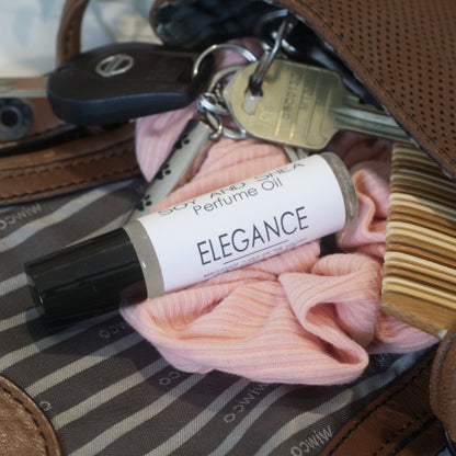 A clear bottle with black lid and white label reading PERFUME OIL ELEGANCE is resting atop an open hand bag filled with keys, pink scrunchie and comb.