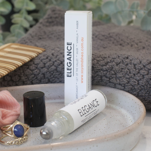 A small narrow clear bottle with the black lid removed and standing to the right revealing a metal roller mechanism rests on a stone tray with rings. Beside the bottle is a white box standing up right with ELEGANCE on the box and bottles label. A grey towel, greenery and wooden comb can be seen in the background.