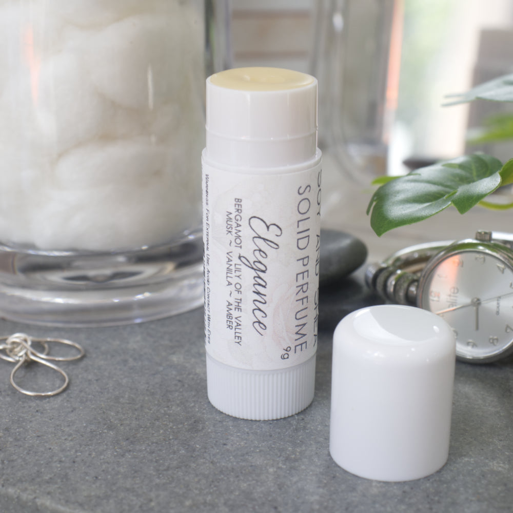 A white wind-up tube stands upright on a bathroom counter top with a silver watch, earrings and greenery.  The lid has been removed and placed to the front right revealing a beige balm.