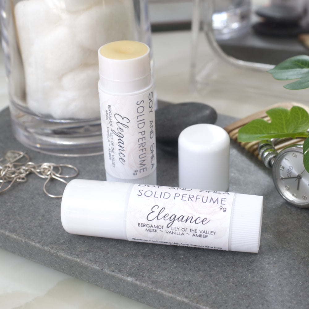 Two white tubes of Solid Perfume are displayed on a grey stone tray along with silver earrings, watch and greenery.   One tube lays flat  displaying the fragrance notes while another tube stands upright behind it with the lid removed and to the right revealing a beige balm