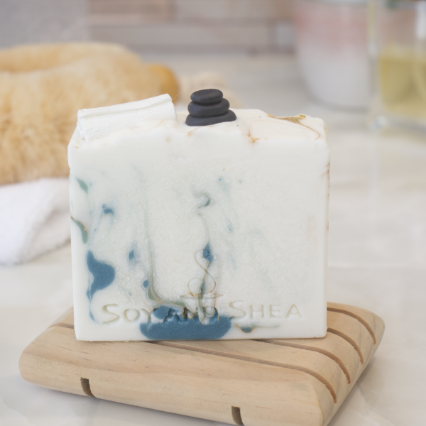 A bar of soap sits on a wooden soap dish and a bathroom counter top with a blurred background.  The soap is off white with dark green and gold swirls.  The top of the soap features soap embellishments of a stack of massage stones and a rolled up white towel.