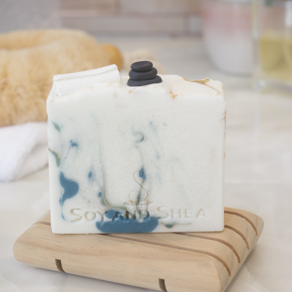 A bar of soap sits on a wooden soap dish and a bathroom counter top with a blurred background.  The soap is off white with dark green and gold swirls.  The top of the soap features soap embellishments of a stack of massage stones and a rolled up white towel.