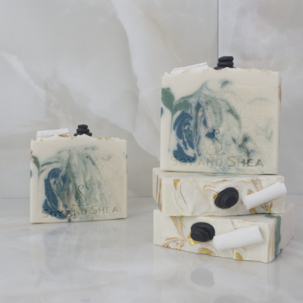 A collection of Elysian Soap bars are displayed against marble tiles.  Two soaps lie flat on the surface stacked up each other showing the top of the soap decorated with soap embellishments of a rolled up towel and a stack of massage stoners.  There are gold swirls through the top of the soap.  On top of these is a third soap standing up to show an off-white soap with green and gold swirls.  A fourth soap stands in the background with the same colours but different design.