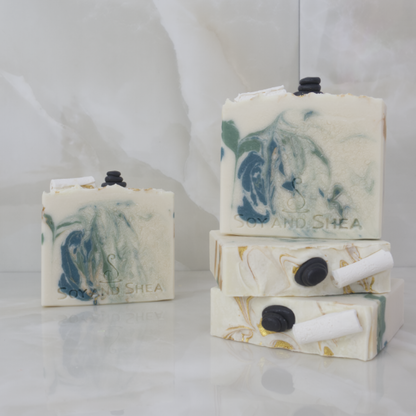 A collection of Elysian Soap bars are displayed against marble tiles.  Two soaps lie flat on the surface stacked up each other showing the top of the soap decorated with soap embellishments of a rolled up towel and a stack of massage stoners.  There are gold swirls through the top of the soap.  On top of these is a third soap standing up to show an off-white soap with green and gold swirls.  A fourth soap stands in the background with the same colours but different design.