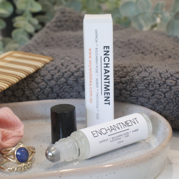 A small narrow clear bottle with the black lid removed and standing to the right revealing a metal roller mechanism rests on a stone tray with rings. Beside the bottle is a white box standing up right with ENCHANTMENT on the box and bottles label. A grey towel, greenery and wooden comb can be seen in the background.