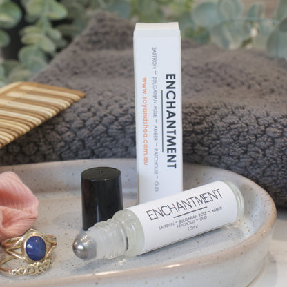 A small narrow clear bottle with the black lid removed and standing to the right revealing a metal roller mechanism rests on a stone tray with rings. Beside the bottle is a white box standing up right with ENCHANTMENT on the box and bottles label. A grey towel, greenery and wooden comb can be seen in the background.
