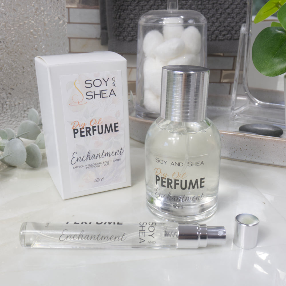 A selection of perfume bottles are presented on a bathroom countertop.  To the front is a clear, narrow, tall bottle labeled as 10ml with a silver cap and behind is a clear round, short bottle with silver lid labelled as 50ml.  Behind sits a white box with label identifying as Dry Oil Perfume Enchantment.