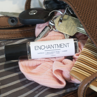 A clear bottle with black lid and white label reading PERFUME OIL ENCHANTMENT is resting atop an open hand bag filled with keys, pink scrunchie and comb.
