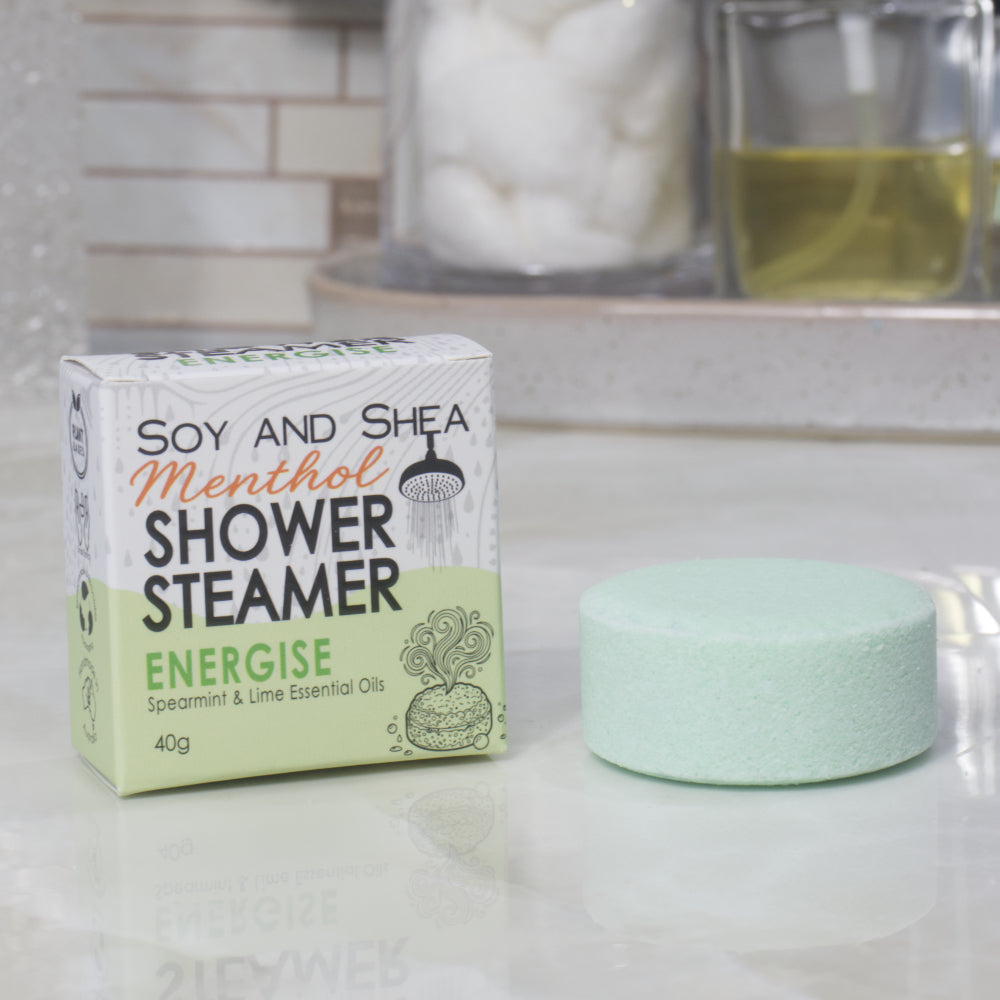 A boxed shower steamer and the steamer itself on a countertop in a bathroom setting. The box reads 'Soy and Shea Menthol Shower Steamer Energise' and the round steamer is a pale green color.